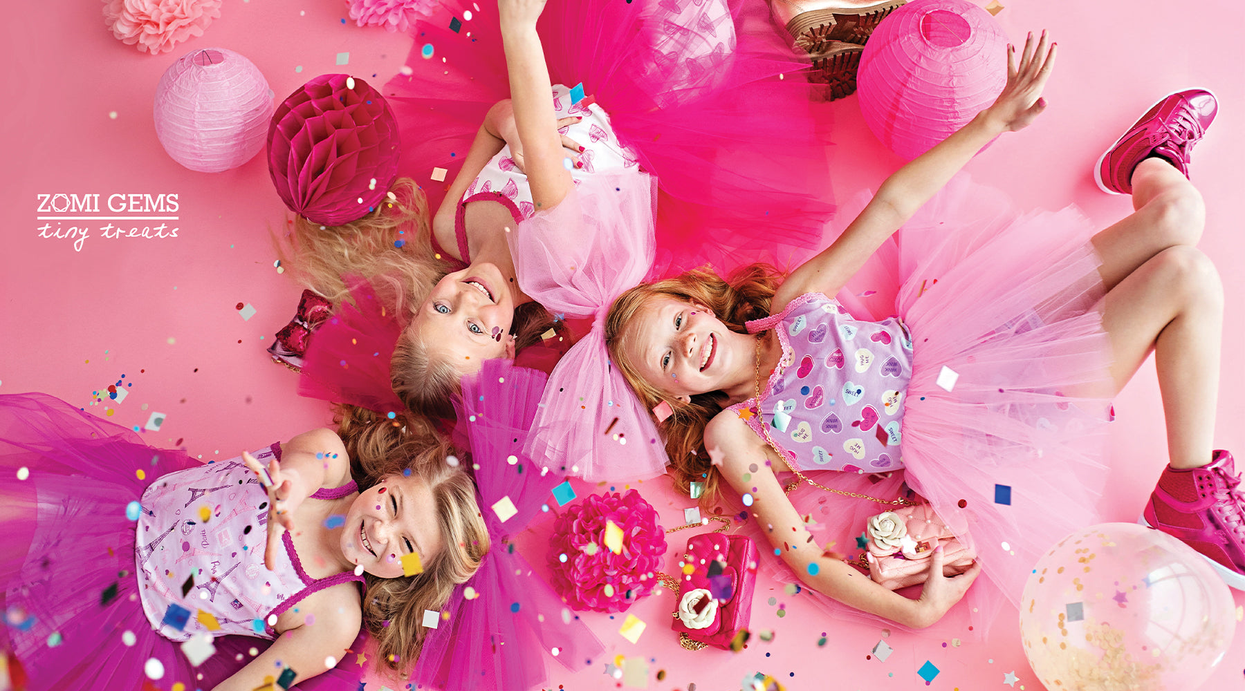 Two girls in pink tutus lying on a pink floor with confetti and Zomi Gems toys.