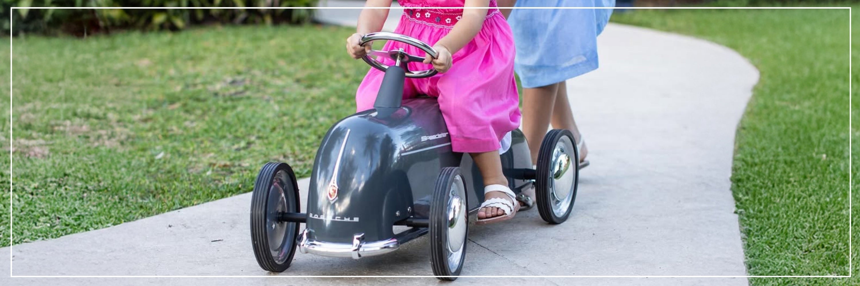 Child in a pink dress riding a toy car on a path with a person pushing from behind.