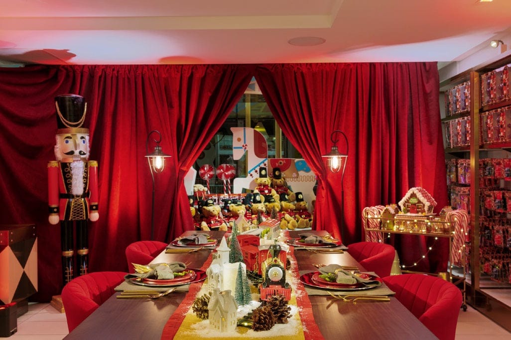 Toy wooden FAO Schwarz soldier in front of a dining room table set with red drapes and a gingerbread house and wooden horse statue.
