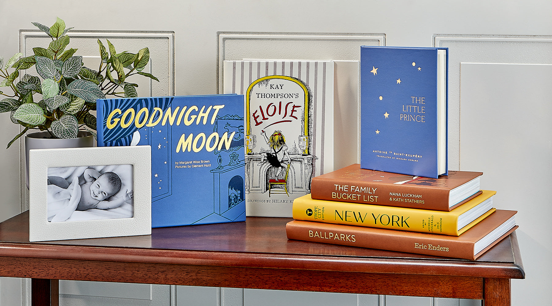 Stack of children's books on a wooden table with a plant and photo frame in the background.