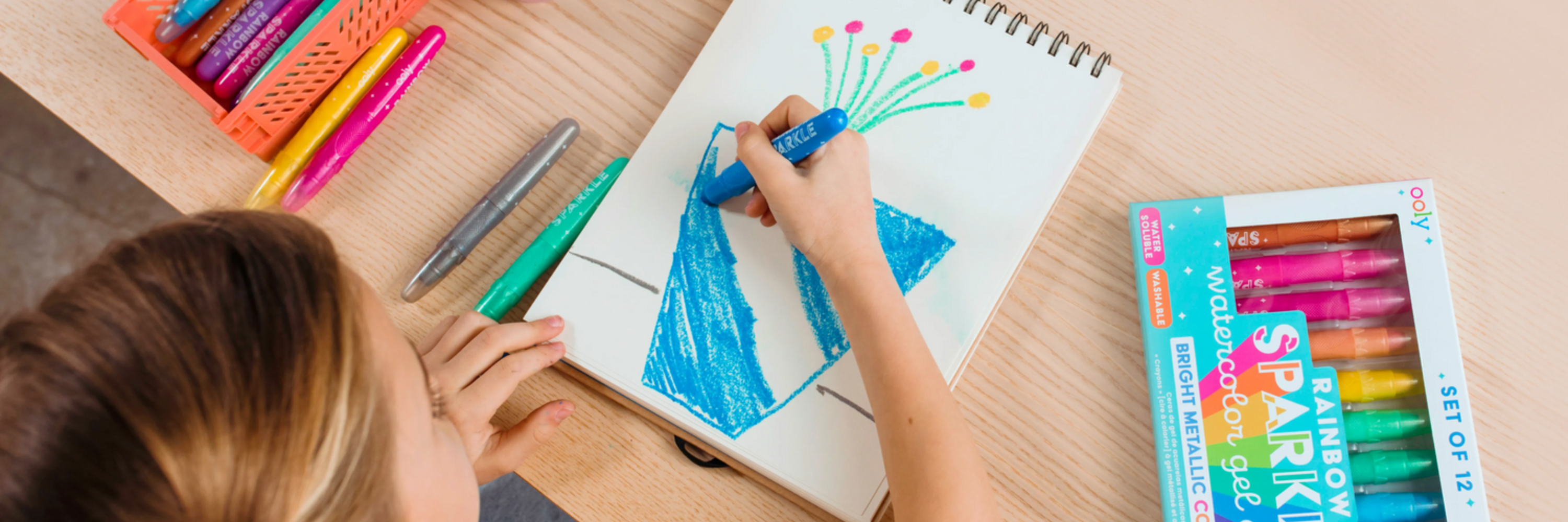 Child coloring with markers on a notebook, surrounded by more markers and a box of markers on a wooden surface.