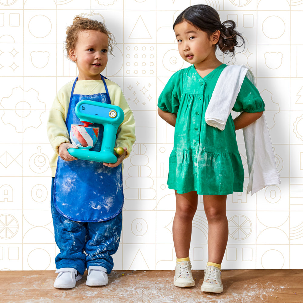 Two children in a kitchen setting, one holding a blue toy mixer and the other with a towel over their shoulder.