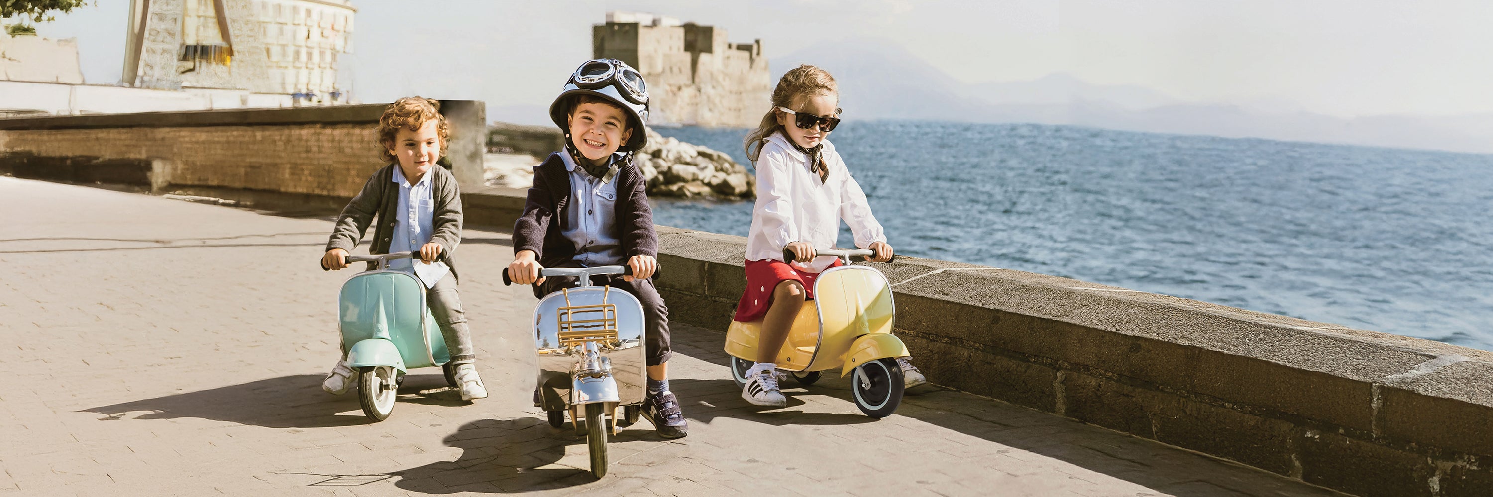 Three children riding small motorcycles by the sea with a castle in the background