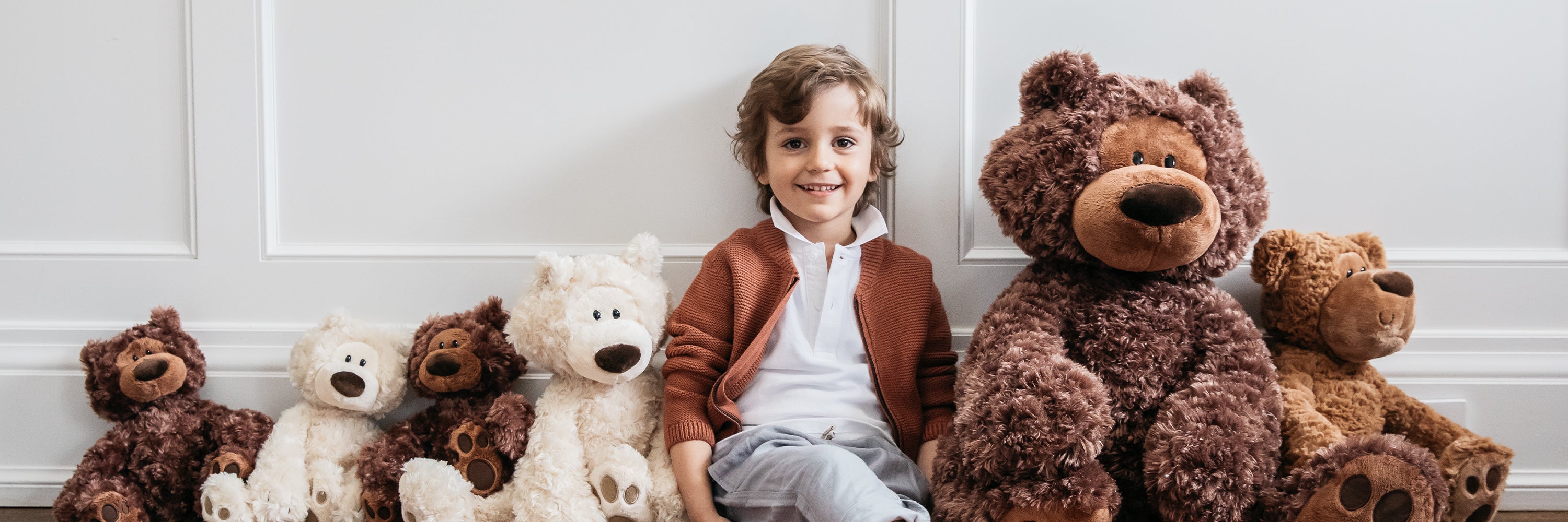 Child sitting with a large teddy bear and other stuffed animals against a white wall.