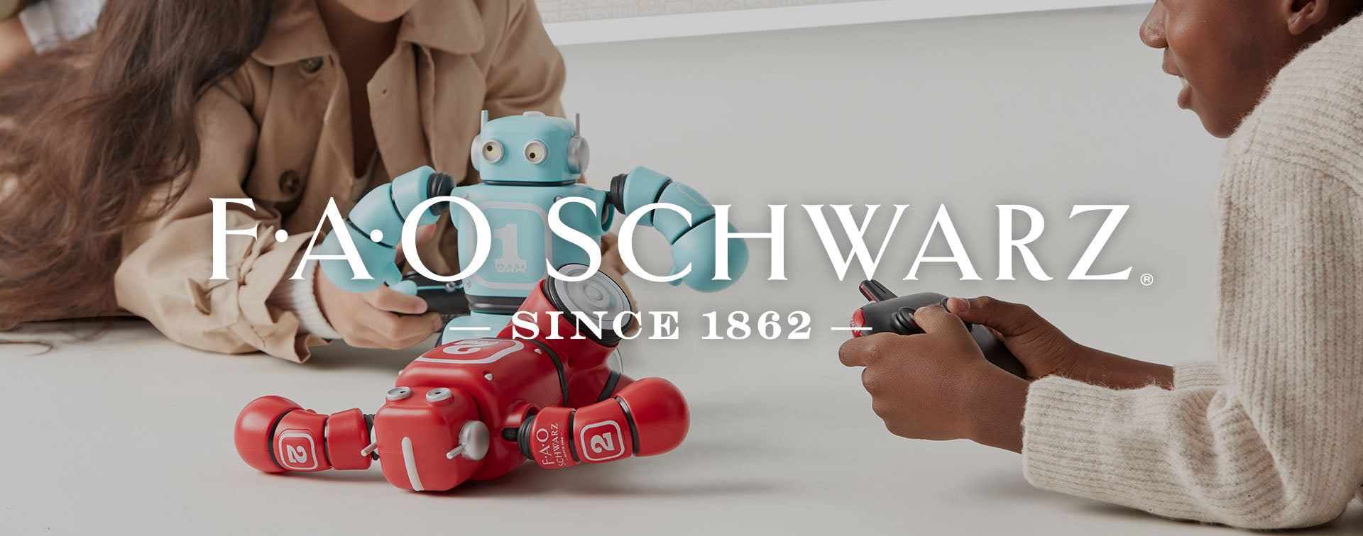 Two people playing with toy robots on a light surface, with 'F.A.O. Schwarz' branding.