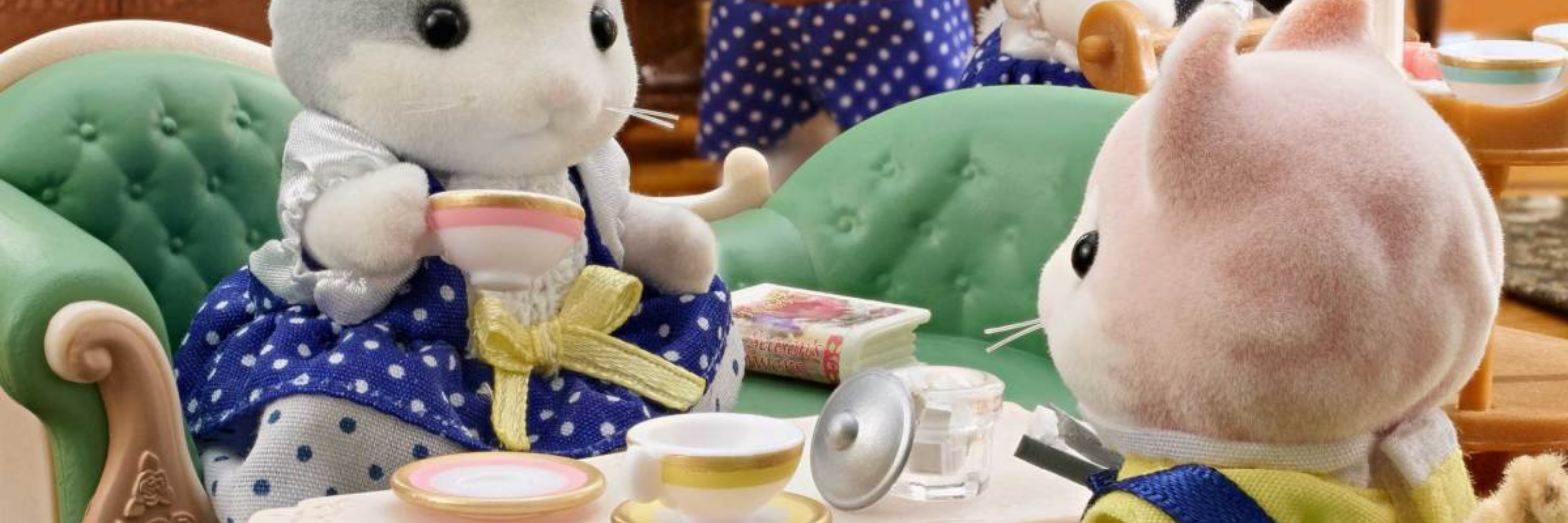 Two plush toys, one in a blue dress with a bow and the other in a yellow outfit, sitting at a table with tea cups and saucers.