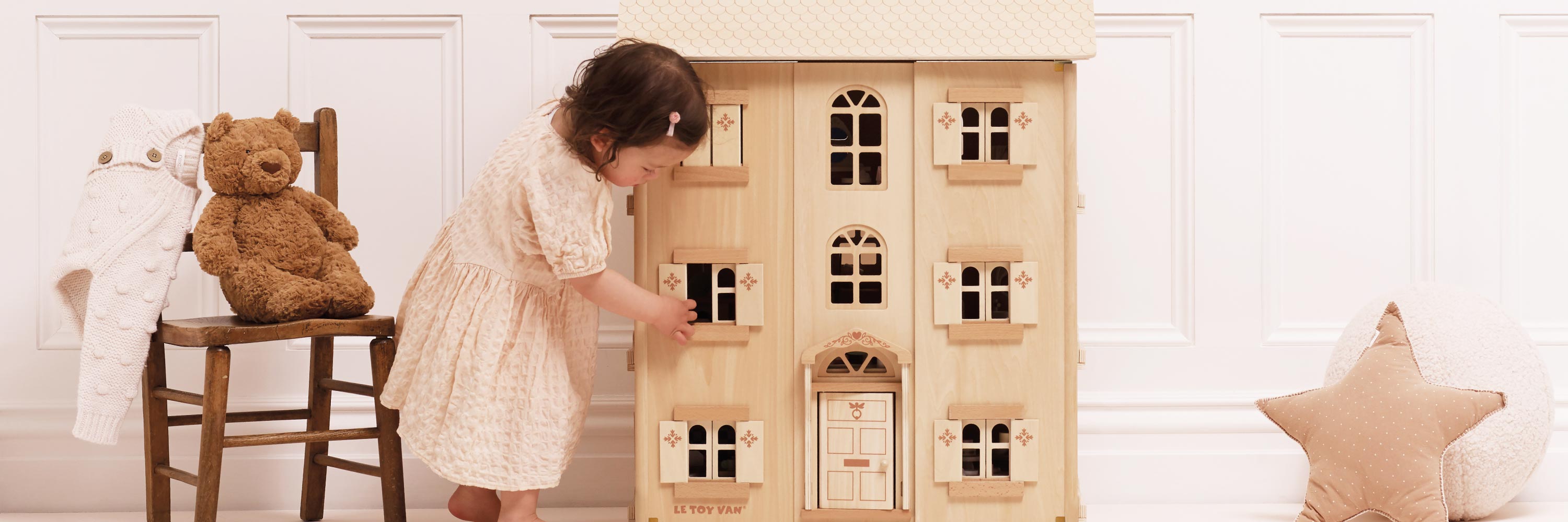 Child playing with a wooden dollhouse in a room with a teddy bear and chair.