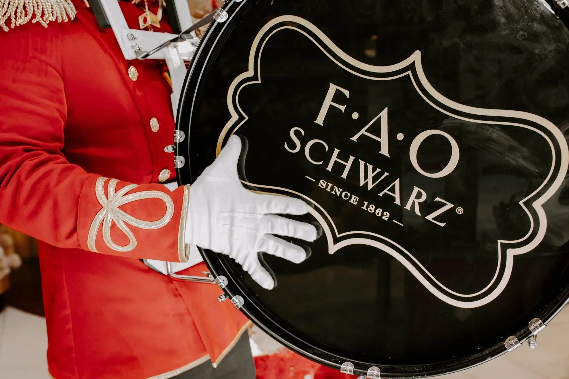 Person holding a large black drum with F.A.O. Schwarz logo, wearing a red uniform and white gloves.