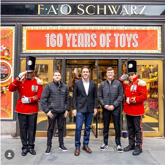 Two men in formal attire standing with two uniformed guards in front of a FAO Schwarz store.