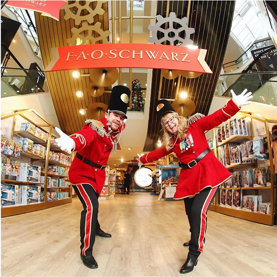 Two people in red uniforms with black pants posing in front of a FAO Schwarz store.